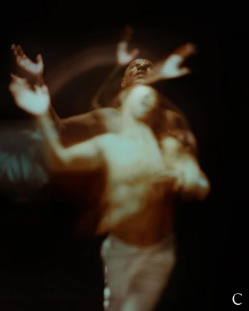 Abstract portrait of blurred figures in motion against a dark background. Dark skin male figure with arms raised. Light leak effect.