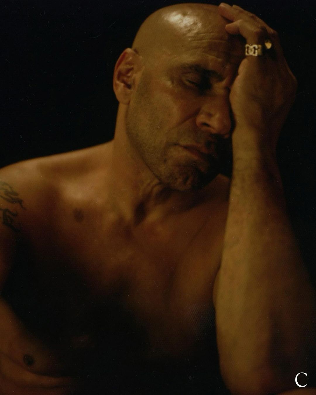 Bald man with hand on face, wearing a ring, set against a dark background in a dimly lit setting with warm tones.