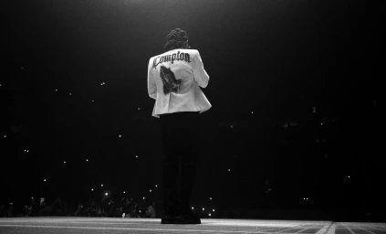 Monochrome rear view of a performer wearing a white jacket with Compton lettering on stage by Fenn O'meally