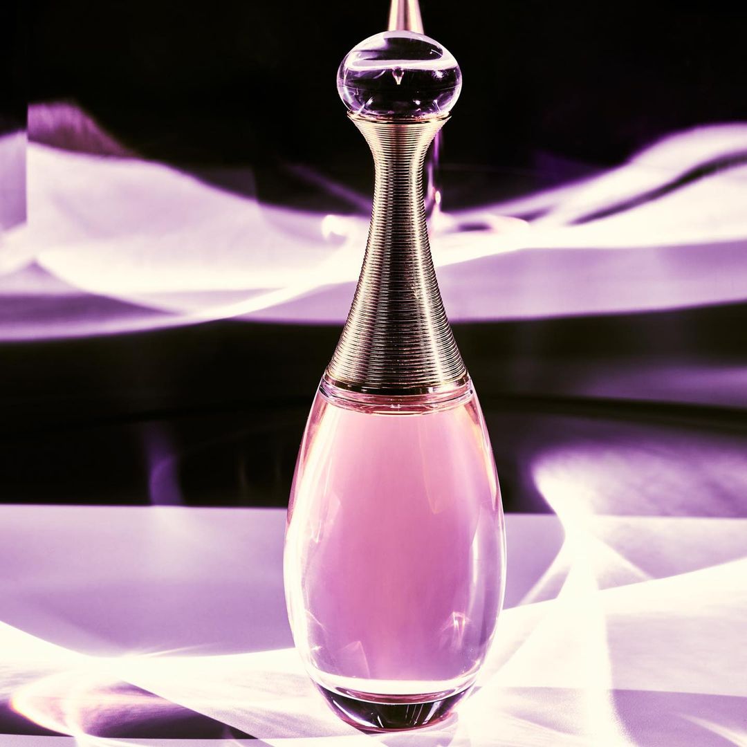 Elegant perfume bottle with gold accents and light pink liquid stands on a reflective surface against a dark, subtly patterned backdrop.