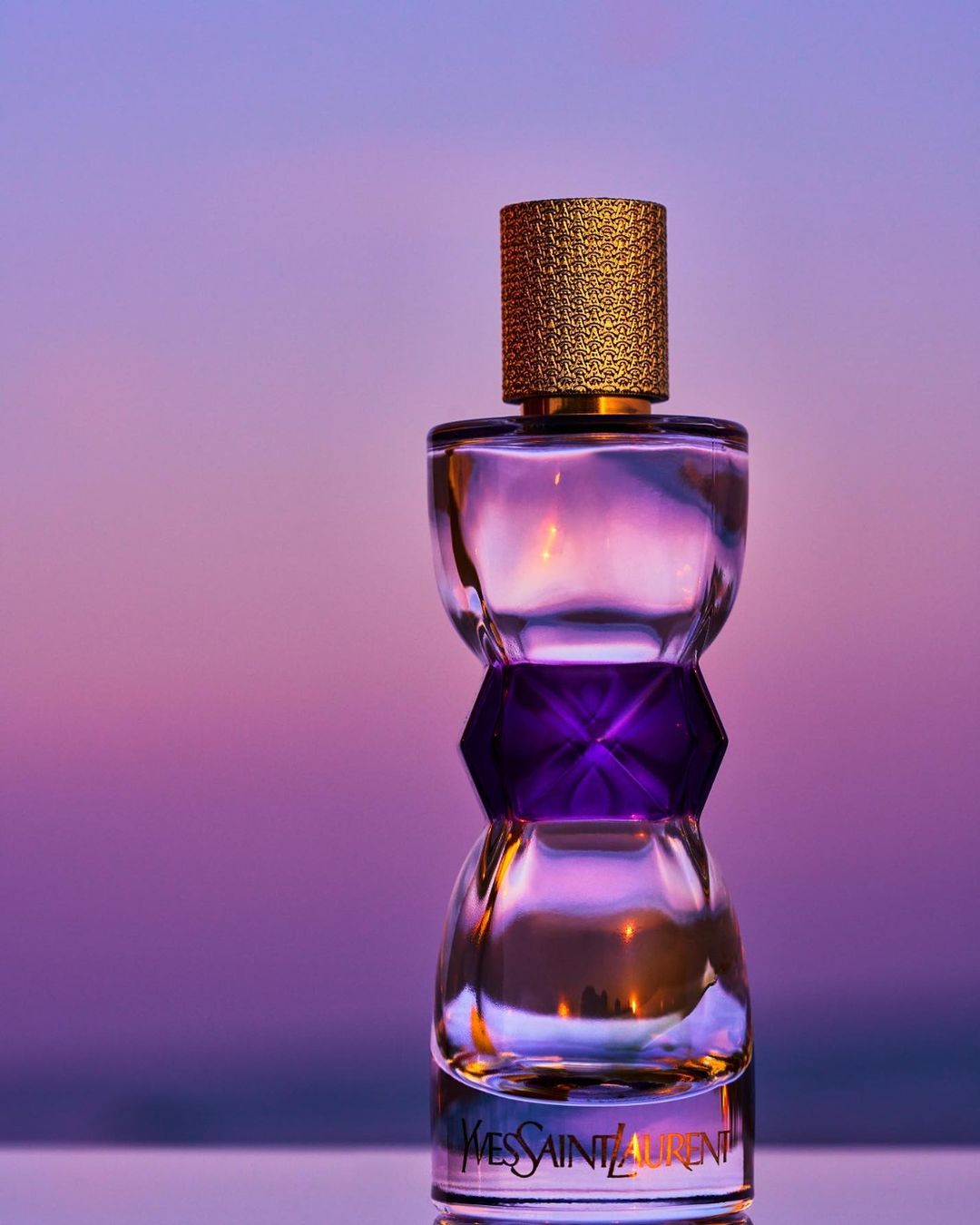 Close-up of a glass Yves Saint Laurent perfume bottle with a textured gold cap against a soft purple background with golden reflections.