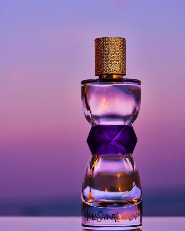 Close-up of a glass Yves Saint Laurent perfume bottle with a textured gold cap against a soft purple background with golden reflections.