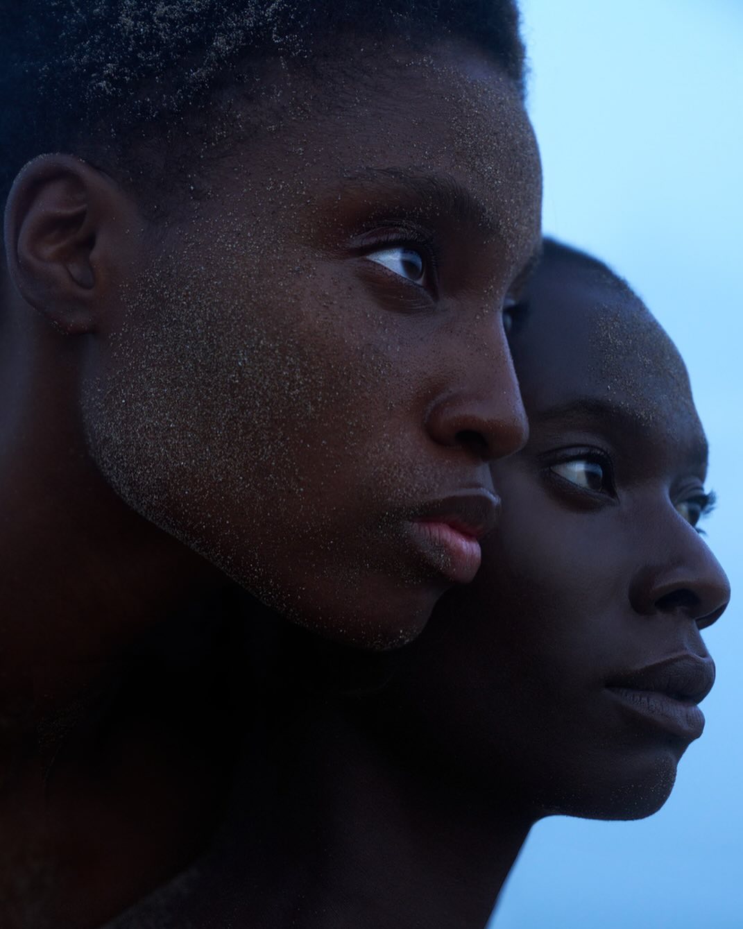 Close-up portrait of two Black models with sand on their skin against an overcast sky, photographed by Fernando Gomez.