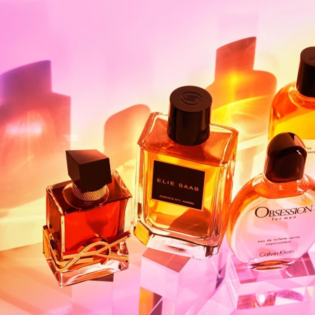 Several glass perfume bottles of various shapes and sizes sit on clear platforms, reflecting light, with a gradient background.