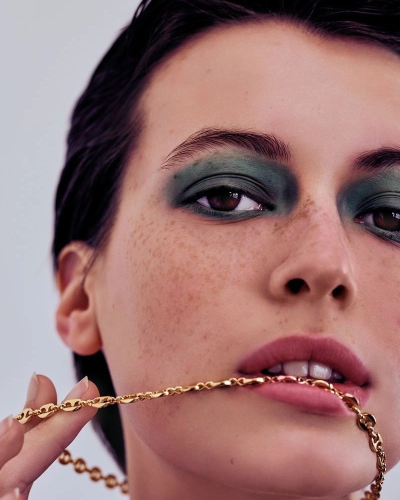 Close-up of a young adult woman with freckles, wearing green eyeshadow and a gold chain necklace, part of it held in her mouth.