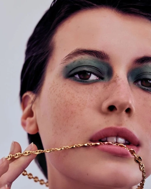 Close-up of a young adult woman with freckles, wearing green eyeshadow and a gold chain necklace, part of it held in her mouth.