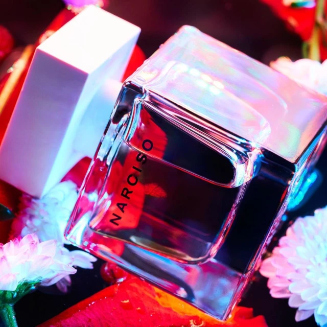 Close-up studio shot of a Narciso Rodriguez perfume bottle with a white cap, surrounded by white and red flowers against a dark background.