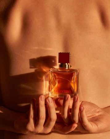 Close-up of hands holding a square Valentino perfume bottle against a bare human back with warm amber lighting.