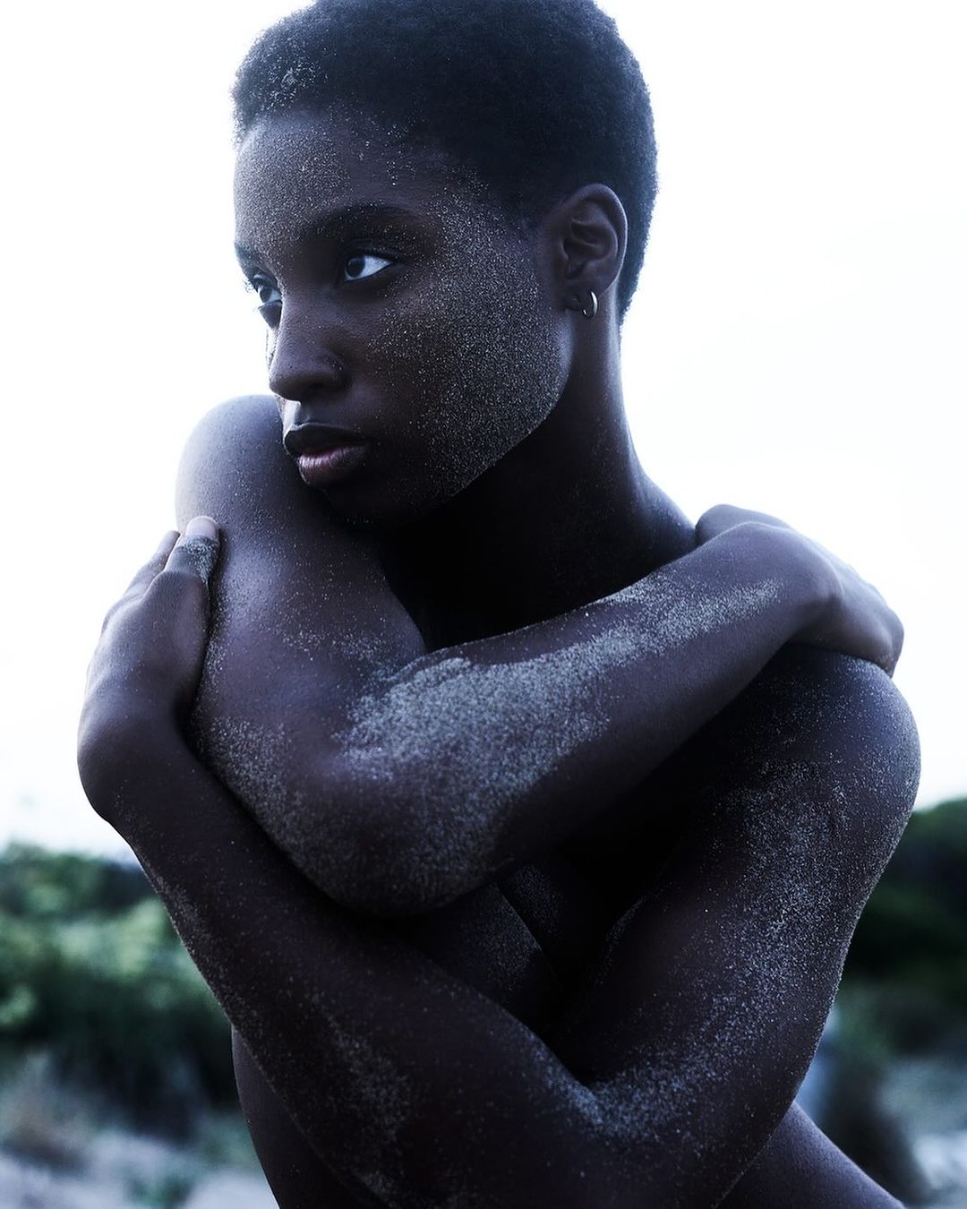 Close-up portrait of a slender, dark-skinned African woman with short hair, arms crossed, body dusted with fine, light-colored particles.