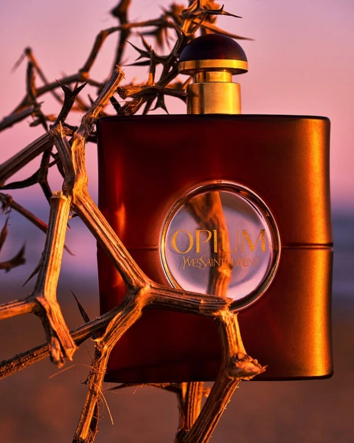 Studio shot of a dark brown Opium Yves Saint Laurent perfume bottle surrounded by thorny branches against a soft pink and purple background.