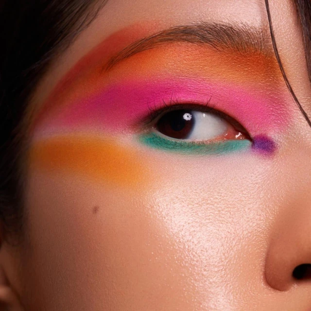 Close-up of a woman's eye with colorful makeup including orange, pink, green, and purple eyeshadow in a studio setting.