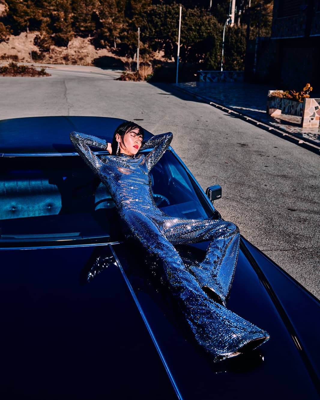 Woman in a shimmering jumpsuit lounges on a dark car hood on a winding road near a hillside with trees under bright sunlight.