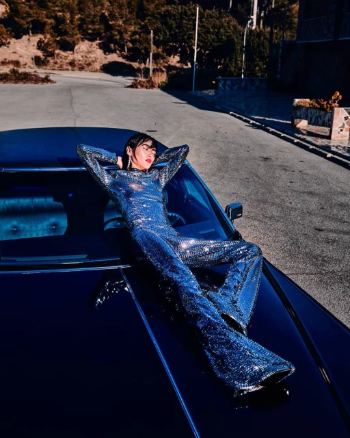 Woman in a shimmering jumpsuit lounges on a dark car hood on a winding road near a hillside with trees under bright sunlight.