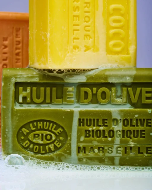 Close-up of stacked olive oil soaps in yellow and green with visible suds and embossed lettering, suggesting a natural skincare product.