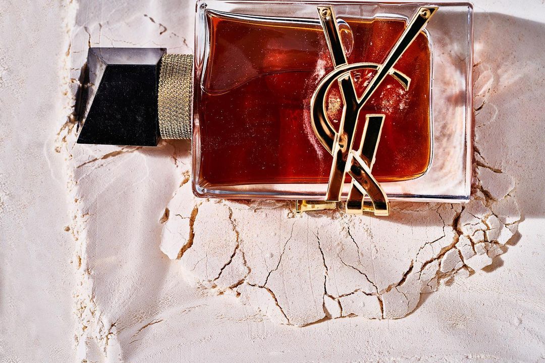 Square perfume bottle with an amber liquid and gold YSL logo is displayed on a textured, cracked, off-white surface, studio lit.