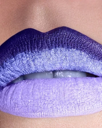 Extreme close-up of a human mouth with lipstick applied in an ombre effect of dark purple to light purple and visible teeth.
