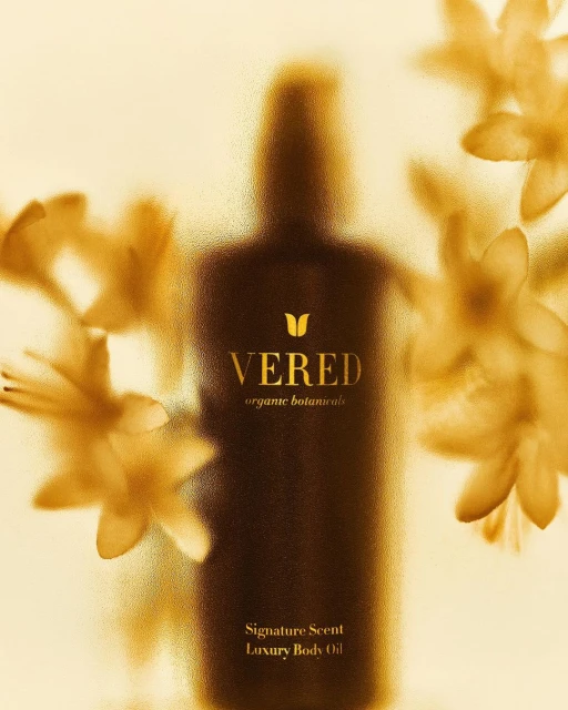 A blurred presentation of a VERED organic botanicals signature scent luxury body oil bottle surrounded by delicate yellow flowers.