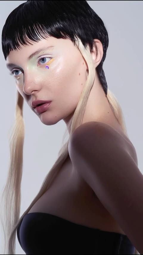 Close-up of a young fair-skinned woman with a dark blunt bob and light hair extensions posing against a light grey studio backdrop.