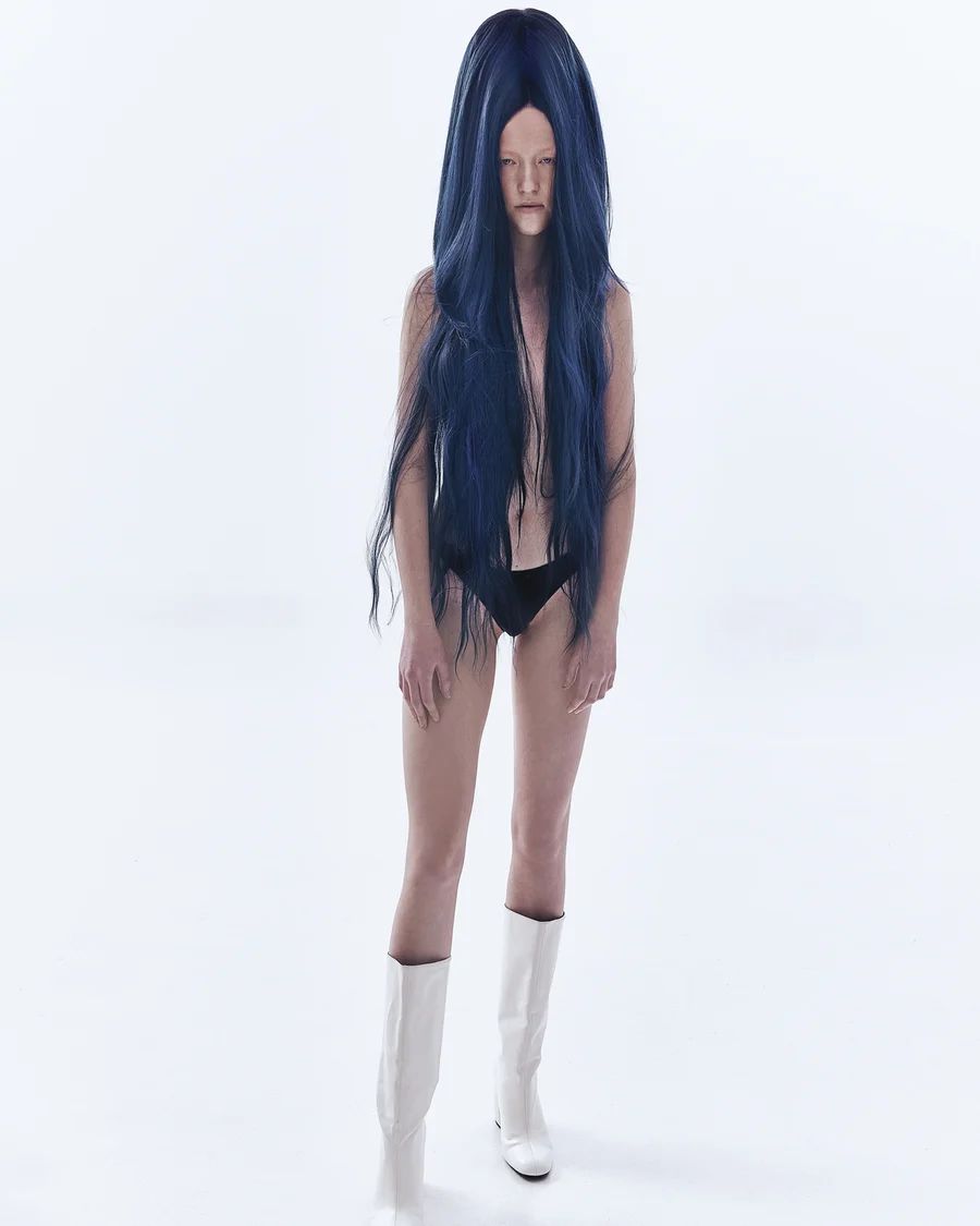 Model with long dark blue wig wearing black briefs and white boots against a plain white background