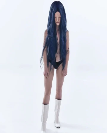 Model with long dark blue wig wearing black briefs and white boots against a plain white background