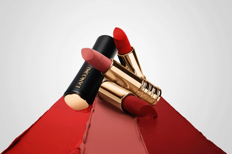 Still life shot of Lancôme lipsticks in black and gold tubes with red, pink and coral colors placed on matching swatches.