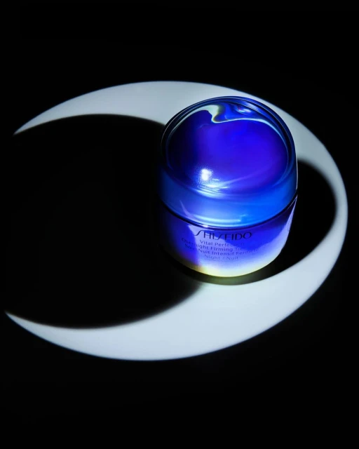 Blue Shiseido Vital Perfection cream jar against a black background with crescent moon lighting, cosmetic product, skincare, studio shot.