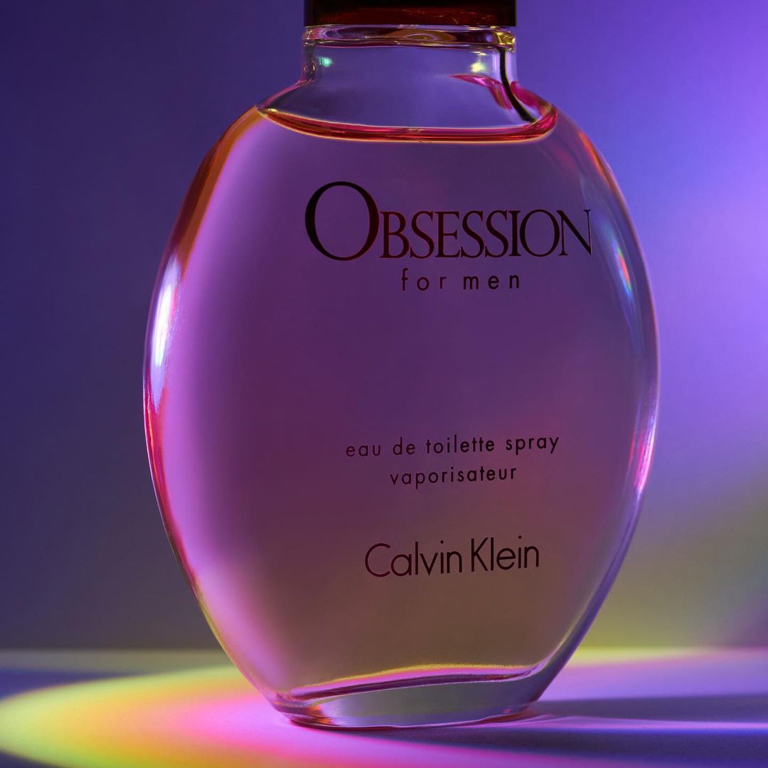 Close-up studio shot of a bottle of Calvin Klein Obsession for Men eau de toilette spray against a softly lit purple and blue background.