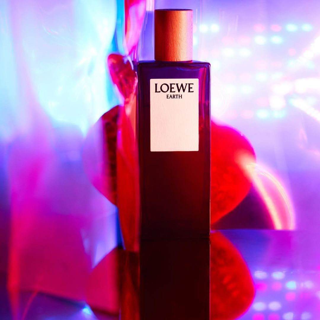 Loewe Earth perfume bottle centered on a reflective surface with a gradient pink and blue background, clean product shot.