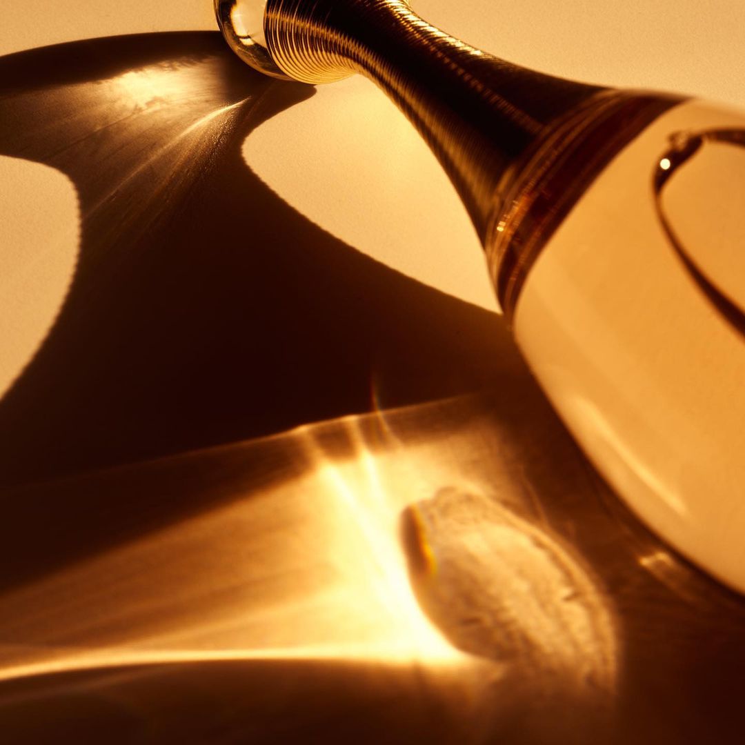 Close-up of a clear glass perfume bottle with a gold accent neck on a reflective surface casting interesting abstract golden light patterns.