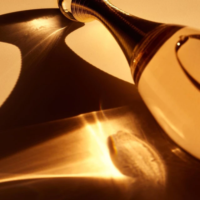 Close-up of a clear glass perfume bottle with a gold accent neck on a reflective surface casting interesting abstract golden light patterns.