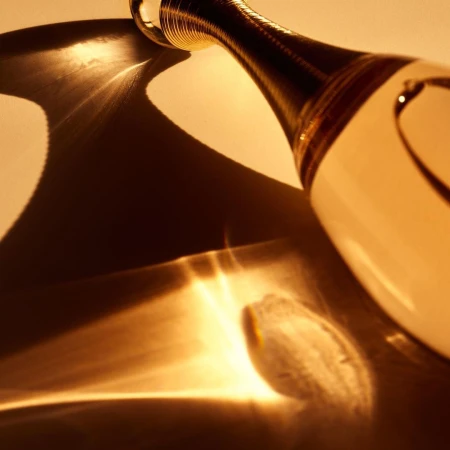 Close-up of a clear glass perfume bottle with a gold accent neck on a reflective surface casting interesting abstract golden light patterns.