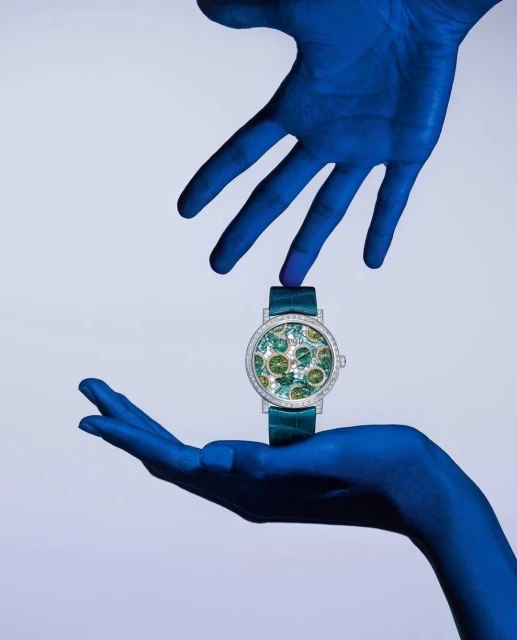 Luxury wristwatch with a green strap and a diamond bezel held by two blue-painted hands against a light blue background in a studio shot.