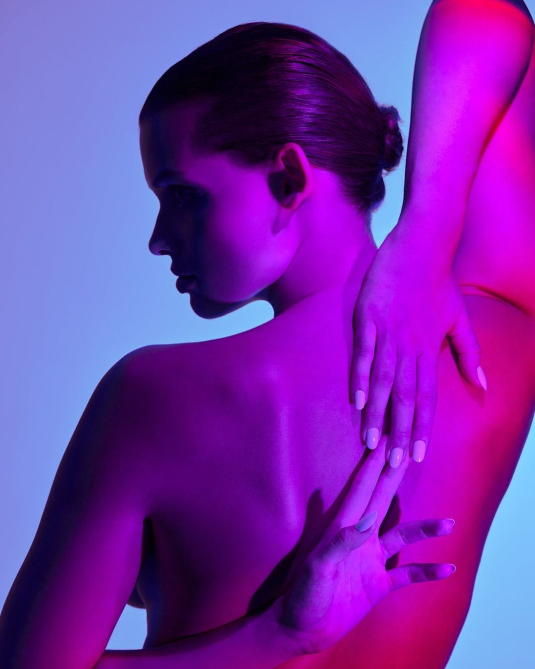 Model with slicked-back hair posing with hands clasped behind back under vibrant magenta and blue lighting by Fernando Gomez.