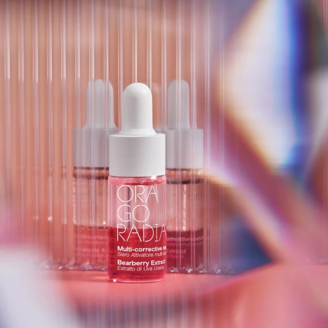 Close-up of Orago Radia serum bottles against a pink-toned background with light refraction effects, highlighting the skincare product's packaging.