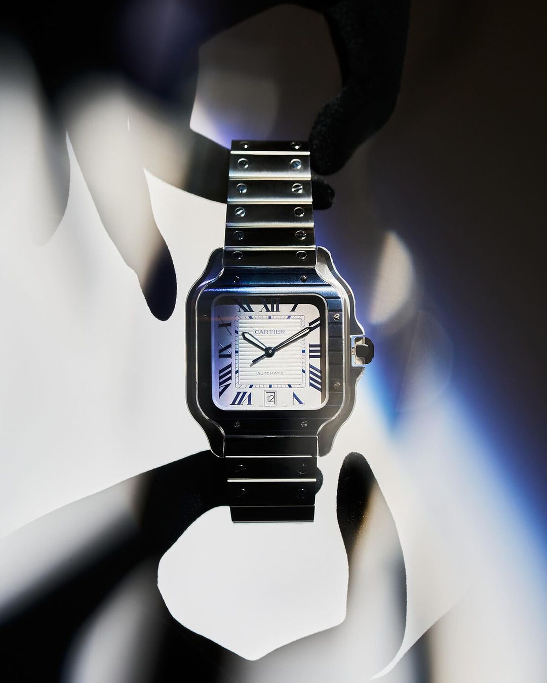 Silver Cartier Santos wristwatch is showcased with dark shadows and a bright, white face. Luxury watch with silver band is set against a light background.