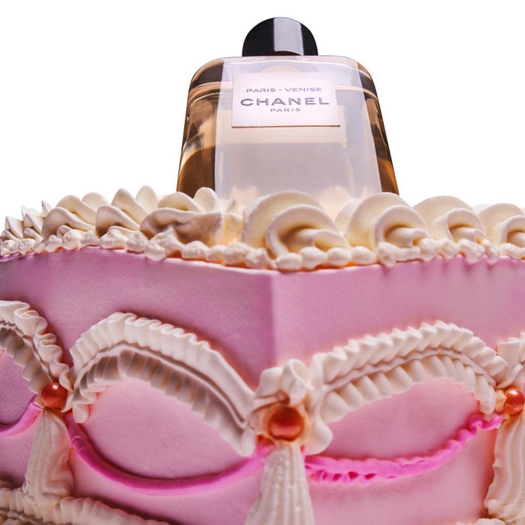 Chanel Paris-Venise perfume bottle displayed on a pink and white cake with decorative frosting details, isolated on a white background.
