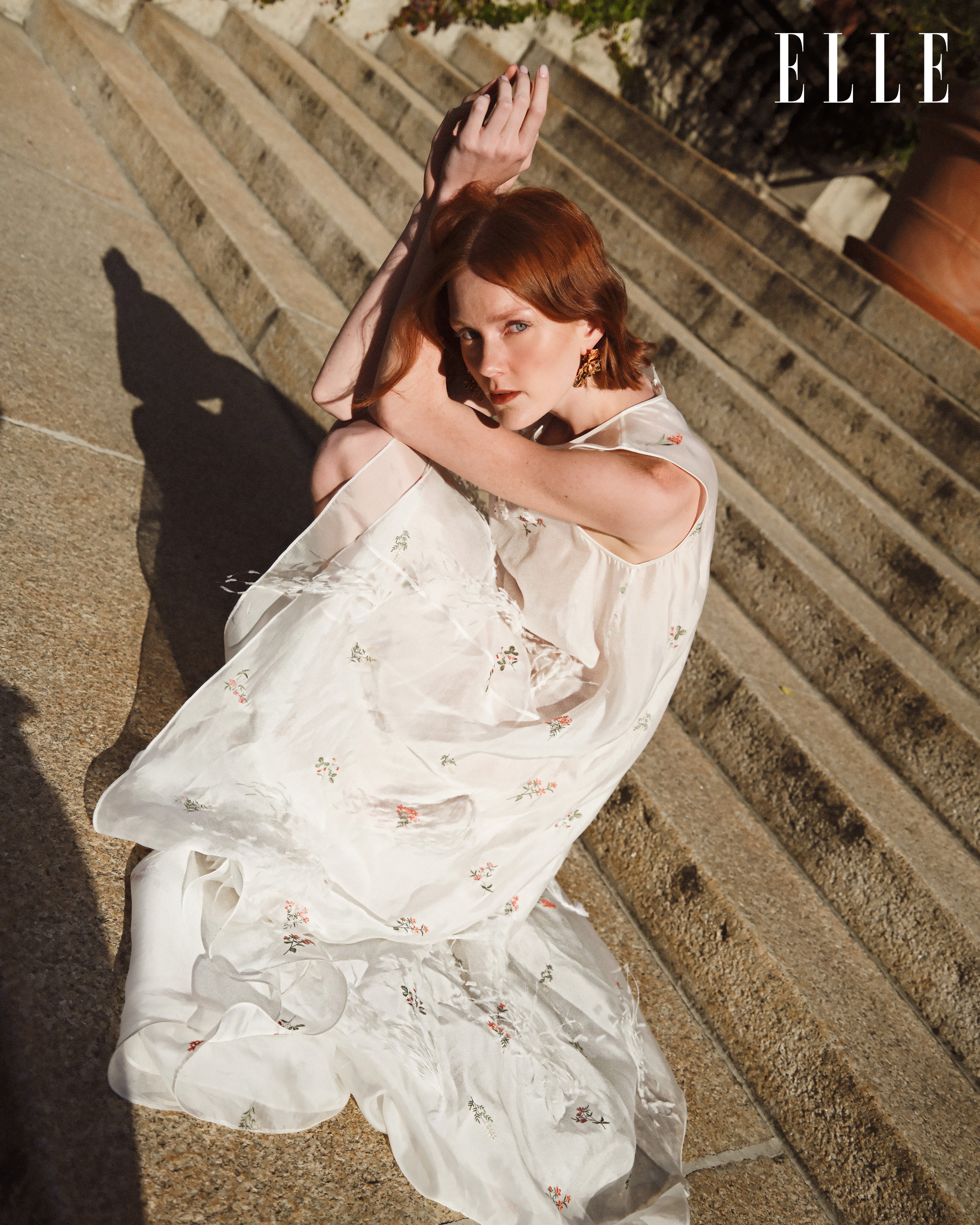 A woman with red hair poses on sunlit stone steps wearing a white floral-patterned dress; elle magazine logo visible in the background.