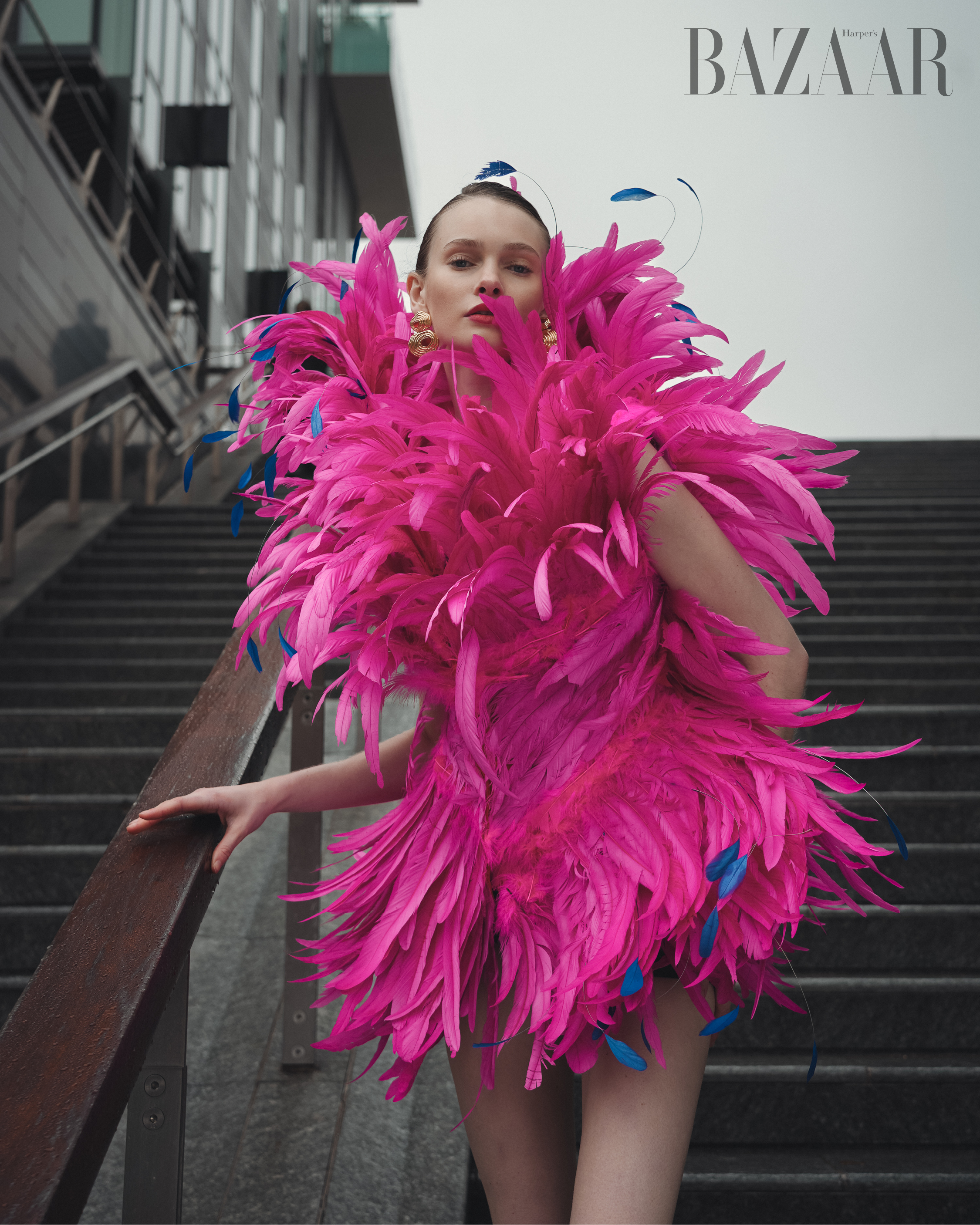 Fashion editorial shot of a woman in a bright pink feather dress posing on outdoor stairs against a modern building, with the Bazaar logo.
