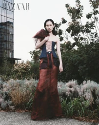 Model posing outdoors in a denim lace-up corset top and a sheer rust-toned maxi skirt beside garden shrubbery and a modern building.