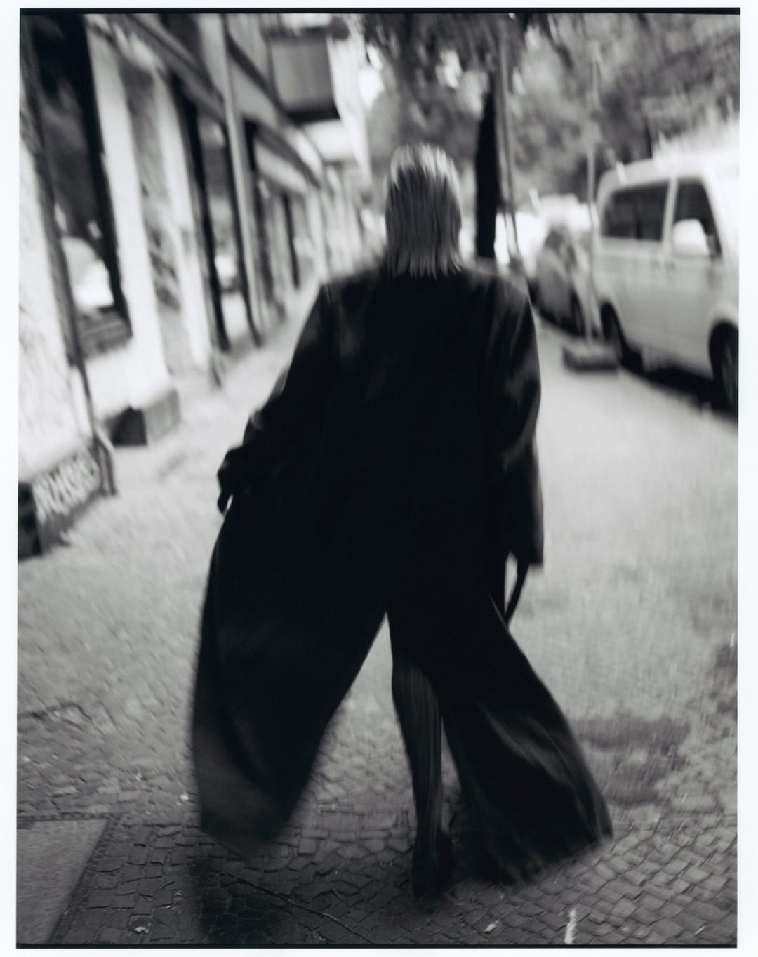 A person walks on a sidewalk in a black-and-white street scene; the figure is wearing a long coat and is seen from the back.