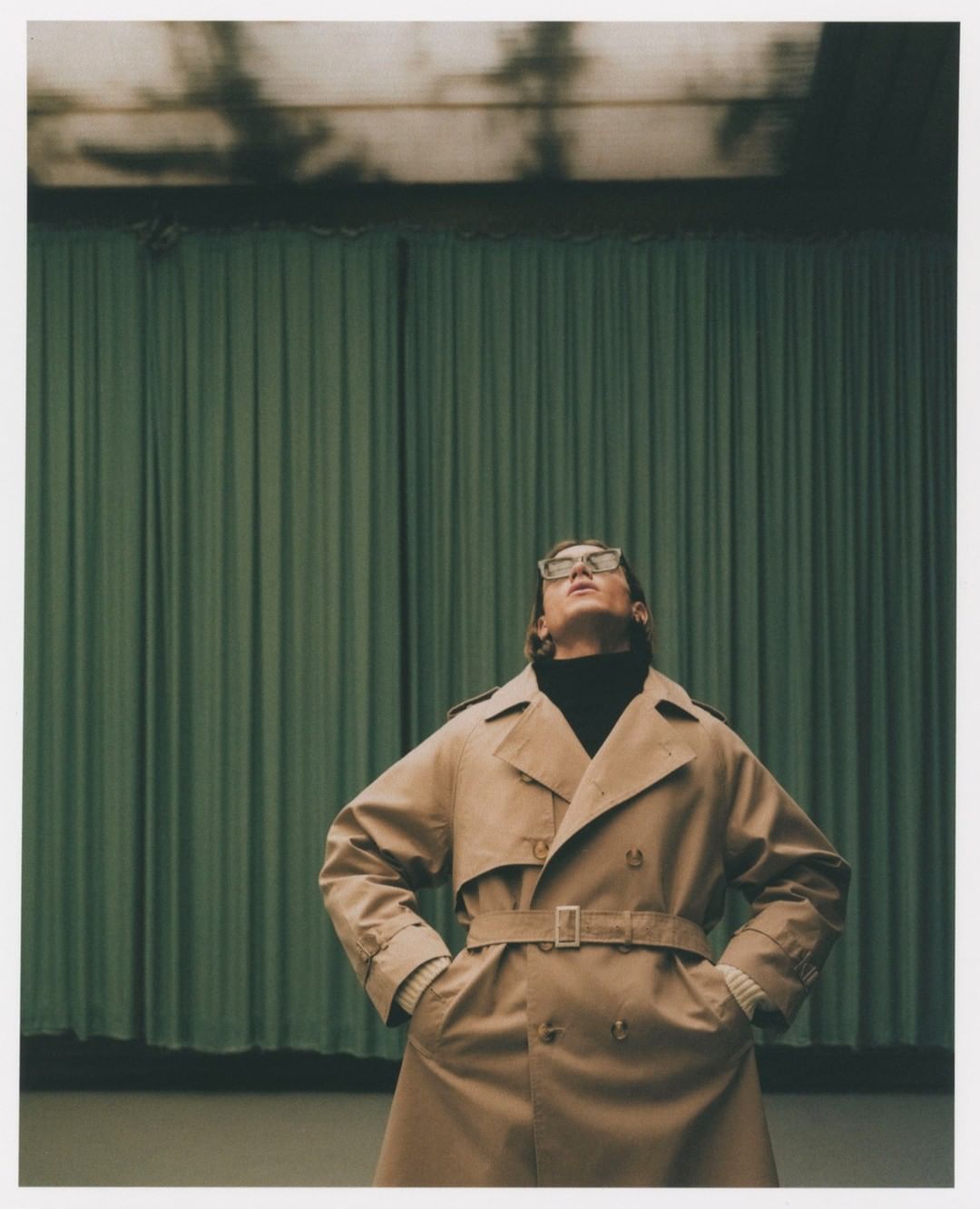 Adult man wearing a trench coat and glasses looks up toward the light in an indoor setting with a curtain in the background.