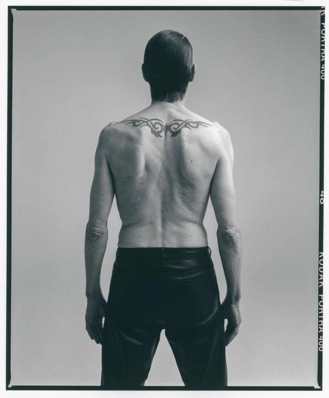 Back view of a shirtless adult male with a tribal tattoo on his upper back, wearing black pants against a grey background, studio lighting.