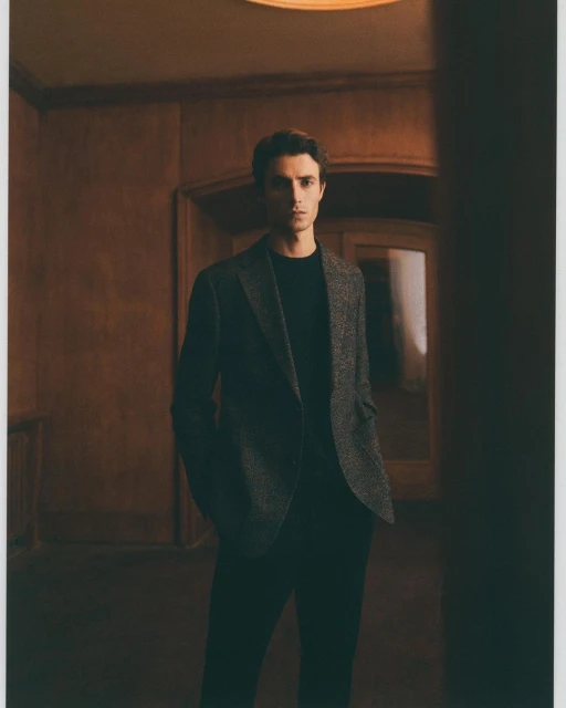 A man in a blazer and dark shirt stands in a dimly lit corridor with panelled walls, under a bright ceiling light.
