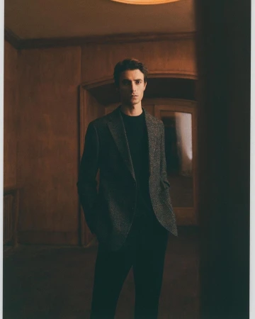 A man in a blazer and dark shirt stands in a dimly lit corridor with panelled walls, under a bright ceiling light.