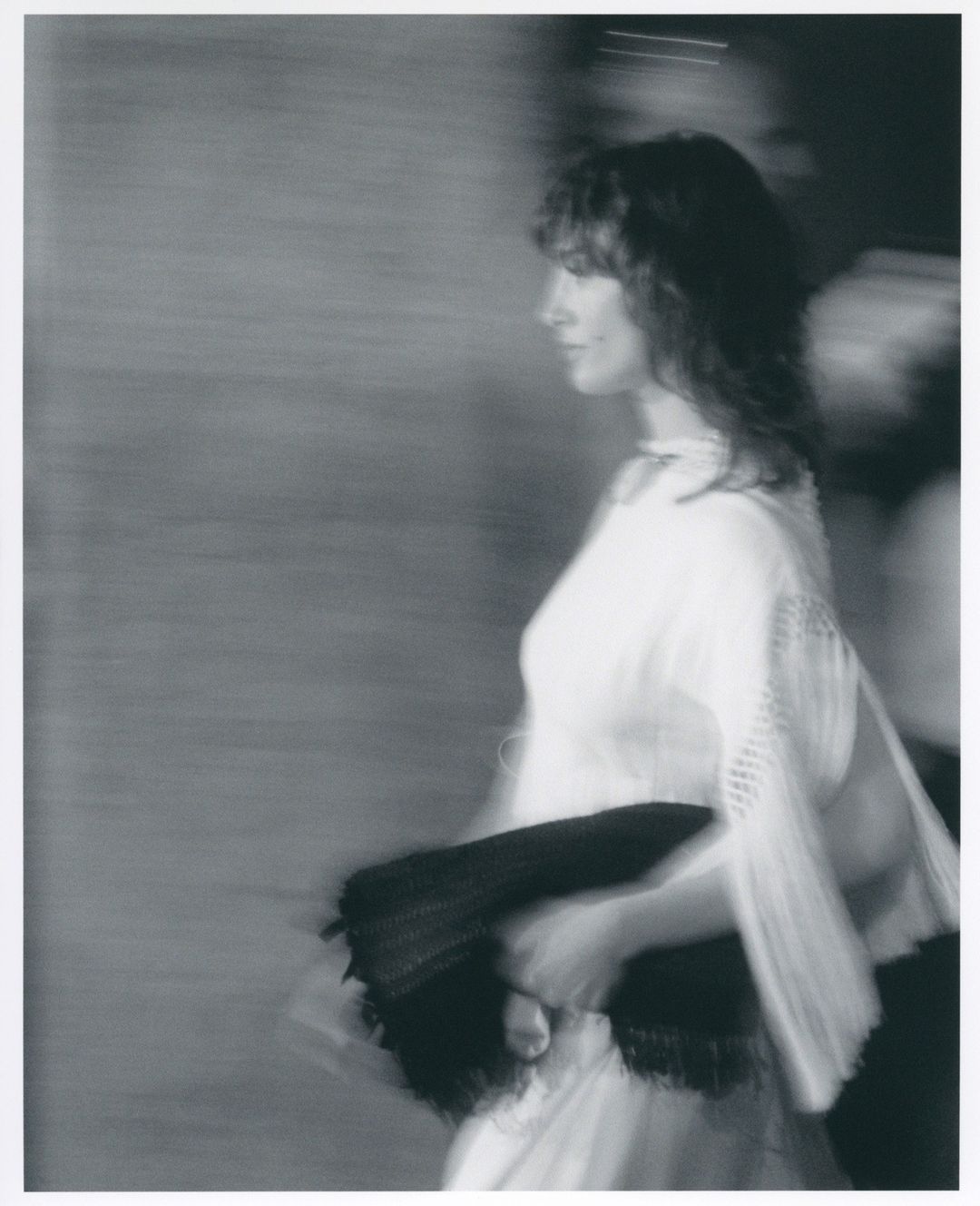Monochrome image of a woman in a light dress holding a dark clutch, side profile, motion blur for fashion or artistic effect.