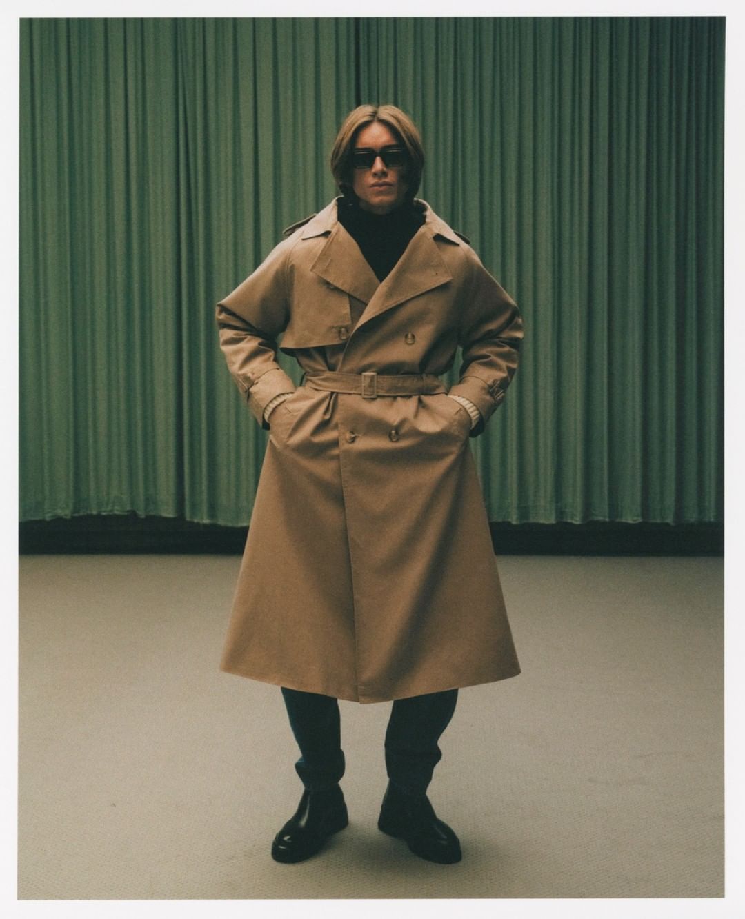 A full shot of a blond adult male in sunglasses, a tan trench coat, and boots standing in front of a green curtain backdrop.