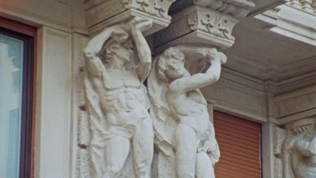 Two sculptures of nude male figures supporting a building structure with stone and window in the background, light colors.