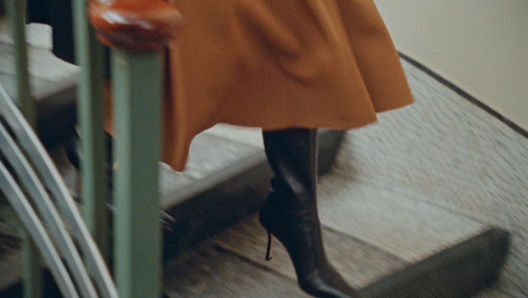 Woman walks down stairs wearing a caramel coat and black, high-heeled boots, with a green railing in the blurred foreground.