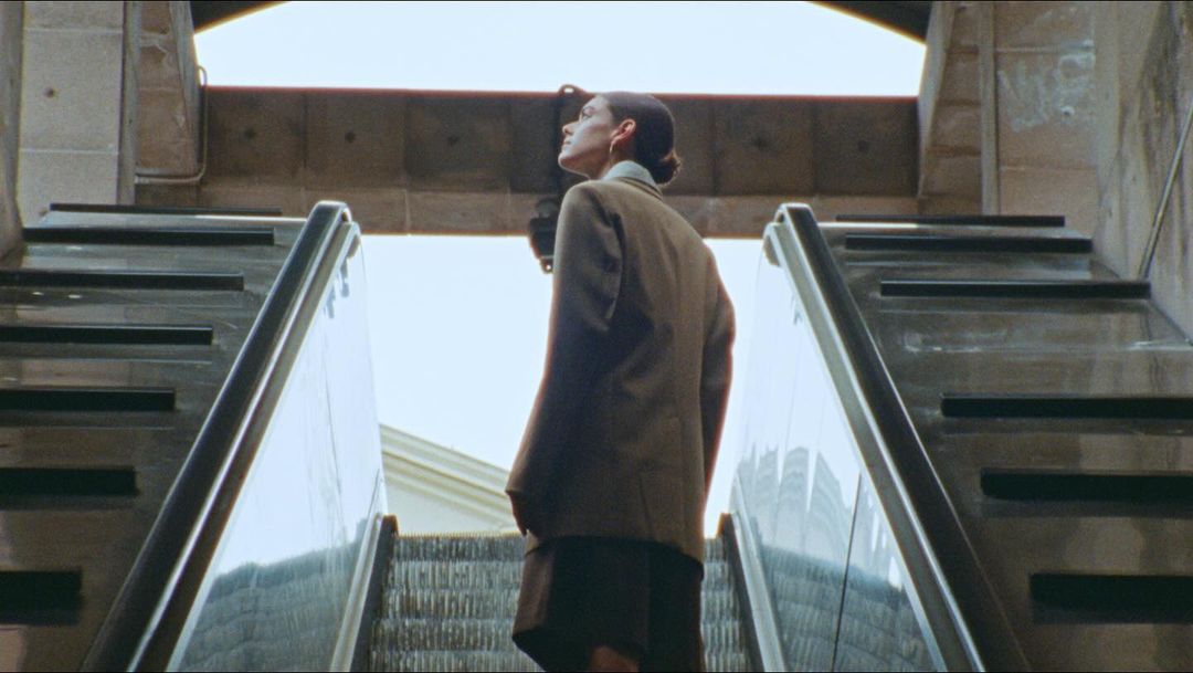 A woman in business attire stands on an ascending escalator with a concrete background, viewed from a low angle, capturing her looking upward.