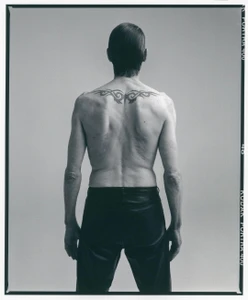 Rear view of a man with dark hair, tribal upper back tattoo, and dark trousers against a neutral grey seamless background.